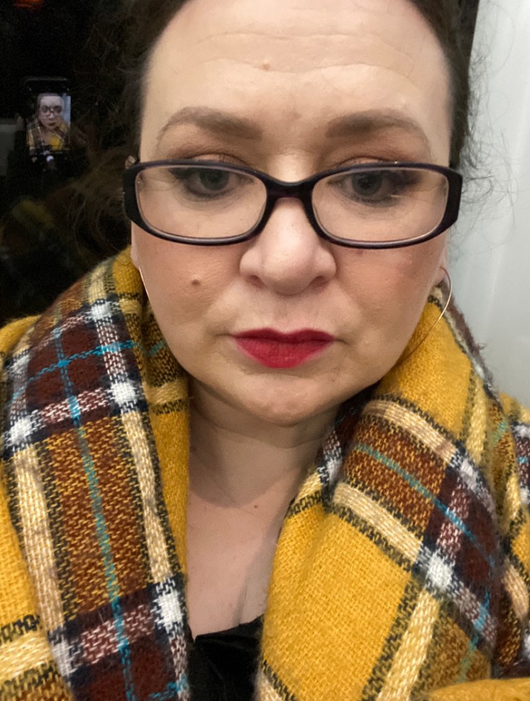 ID: Disabled & chronically ill white cis woman, femme presenting in full makeup with red lipstick. Wears glasses, brown hair tied up off face, beauty spot on left cheek. Wears thin large gold hoop earrings, a mustard, brown, white & teal tartan blanket as shawl. Photo is selfie taken on Dublin train at night. iPhone image of same is reflected behind her in train window. 
